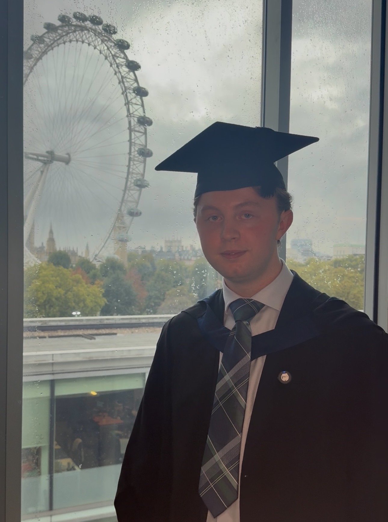 Graduate in cap and gown poses by a window, with the London Eye, rainy skyline, and autumn trees behind him